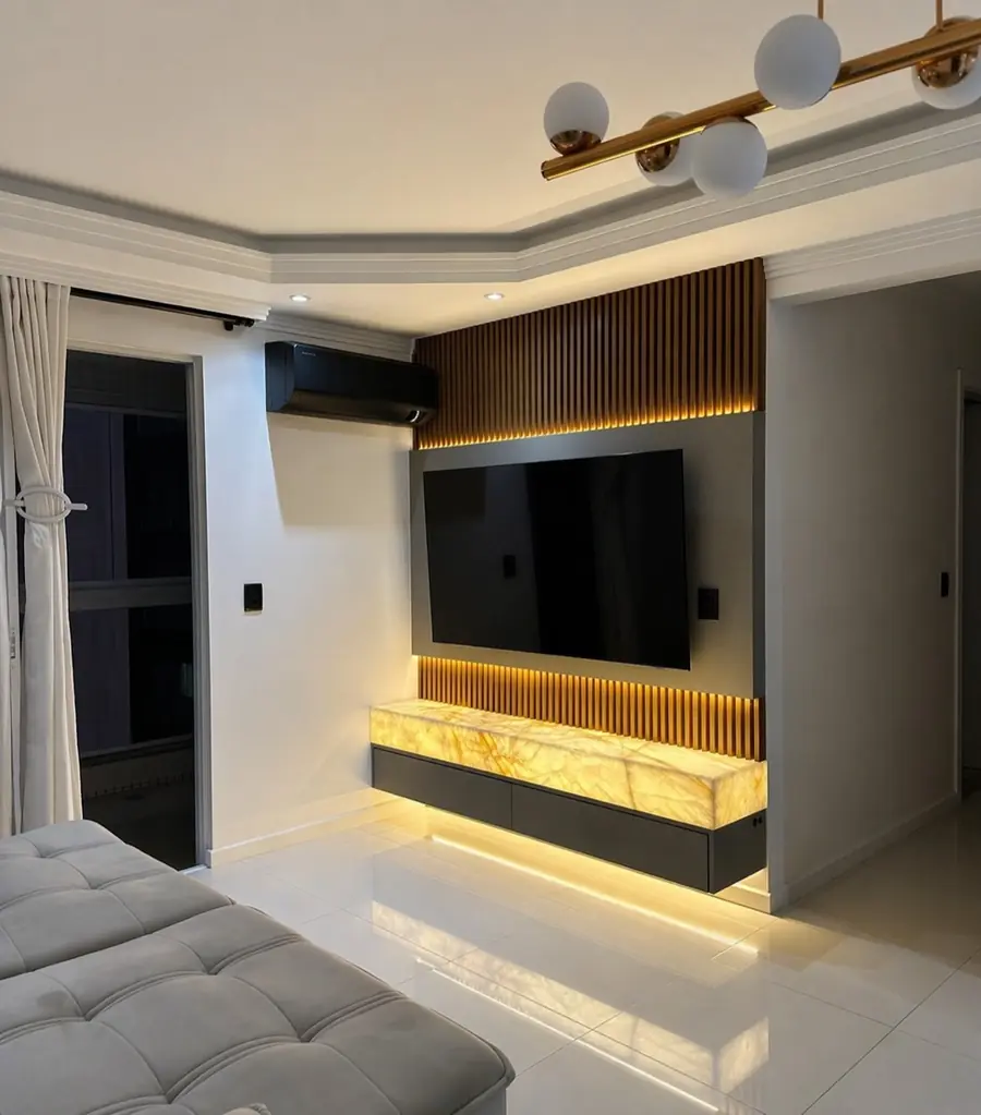 Painel de TV em ripas de madeira, base de mármore âmbar iluminada por LED e luminária de globos dourados no teto.