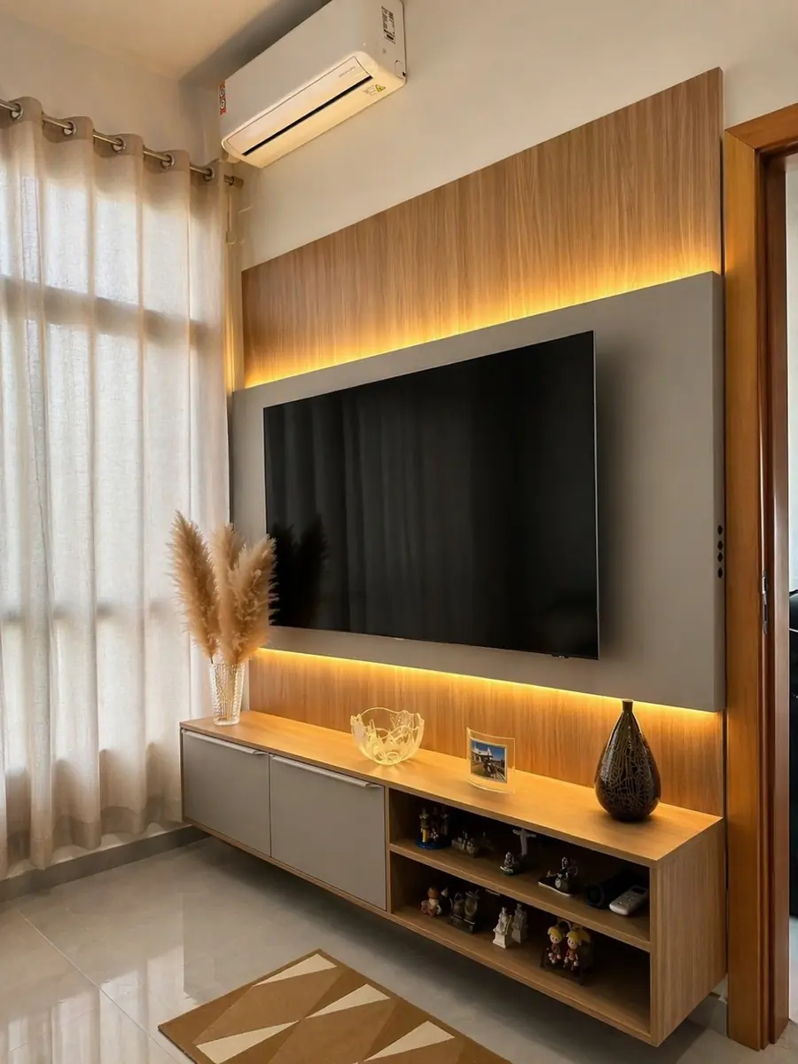 Painel de madeira com TV embutida, iluminação âmbar em LED, nichos com bibelôs e vaso de capim-dos-pampas seco.