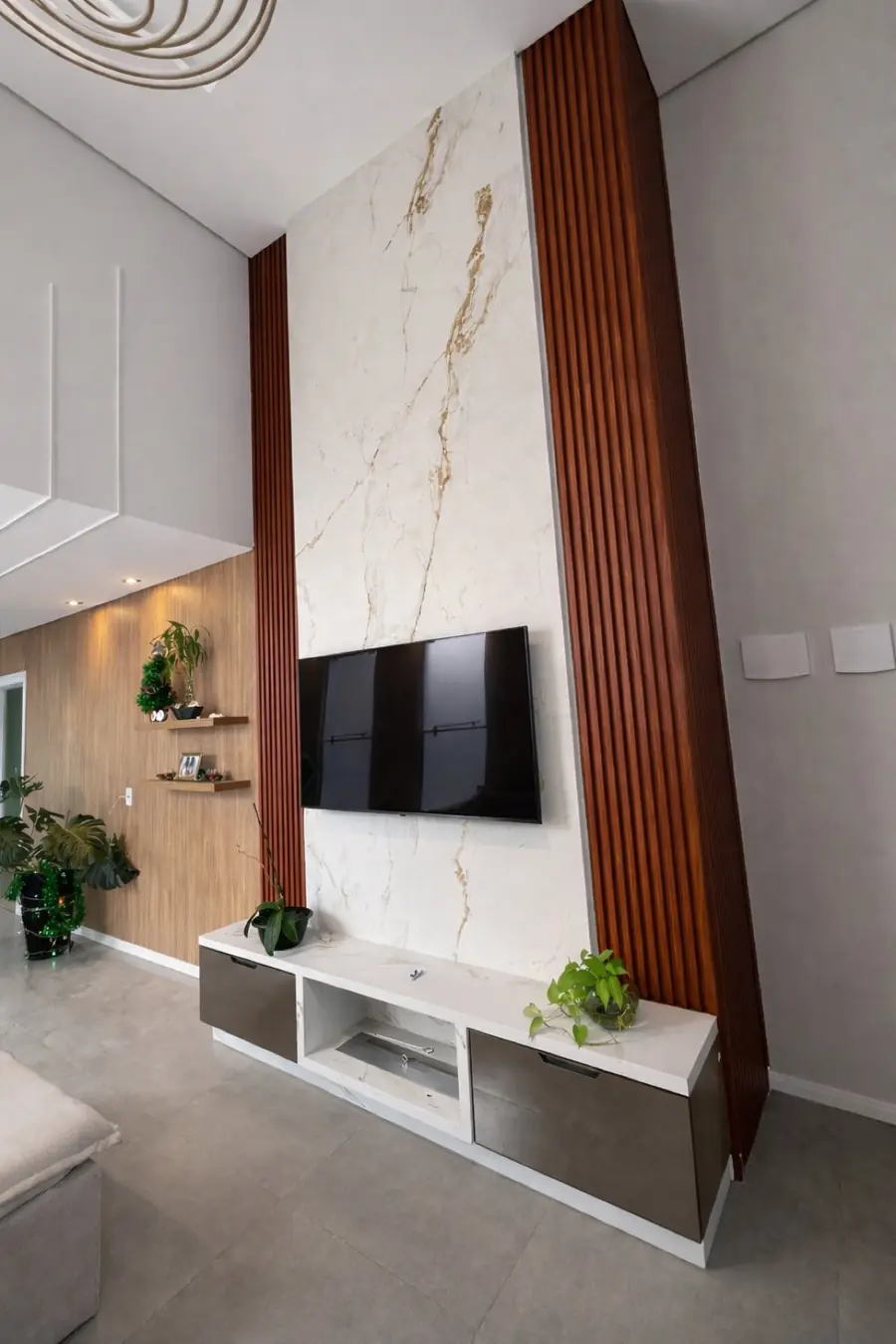 Painel de TV em porcelanato marmorizado ladeado por ripas de madeira avermelhada, do piso ao teto alto.