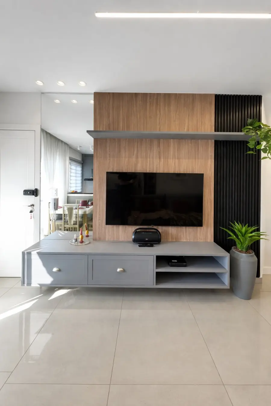 Painel de TV em madeira clara com faixa ripada preta, prateleira suspensa e rack cinza sobre piso porcelanato.