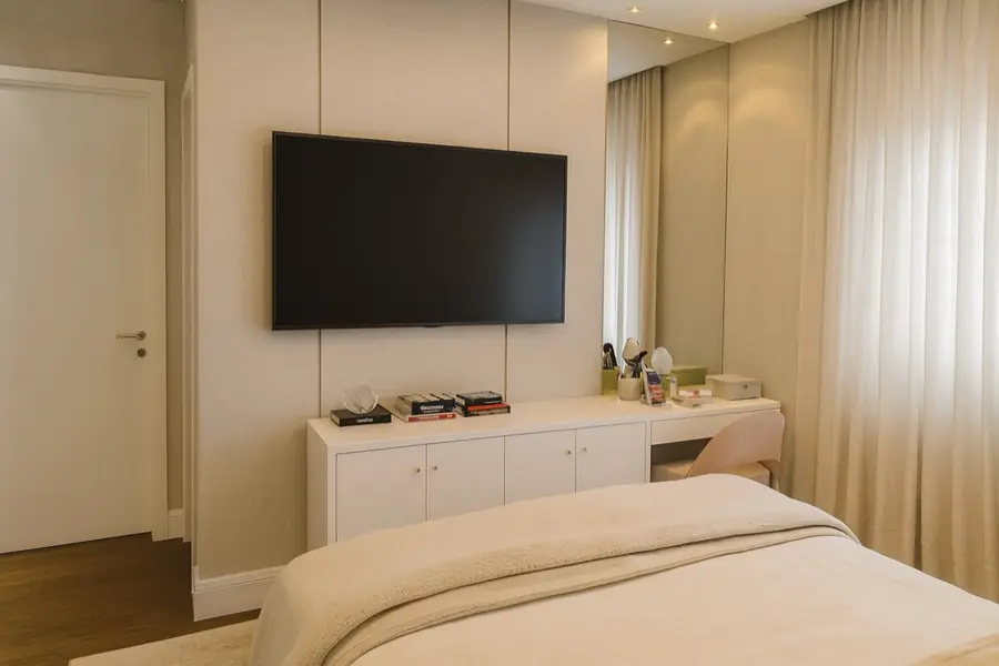 Quarto com painel de madeira clara, TV embutida, aparador branco com gavetas e penteadeira integrada.