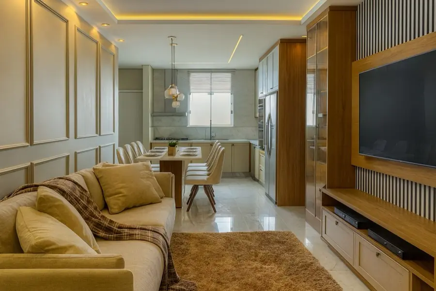 Sala de estar em amarelo mostarda, sofá de canto, tapete peludo e cozinha integrada ao fundo.