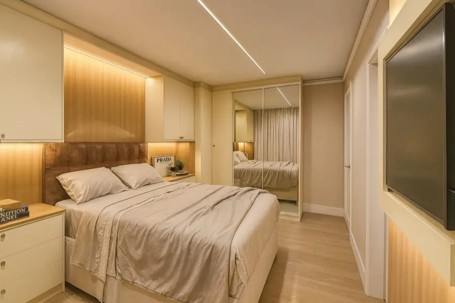 Quarto com cabeceira em courino marrom, armário de portas espelhadas e faixa de luz no teto.